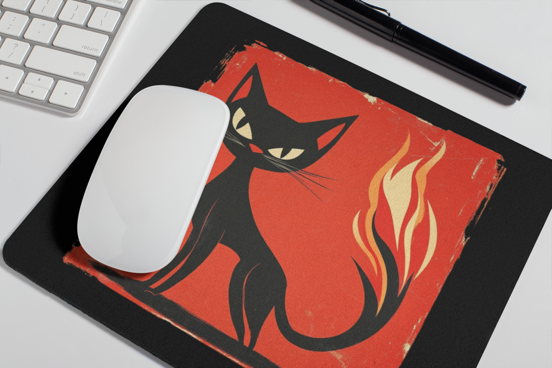 A black mousepad with a red square graphic featuring a black cat and flames, shown on a white desk with a white mouse, part of a keyboard, and a black pen.